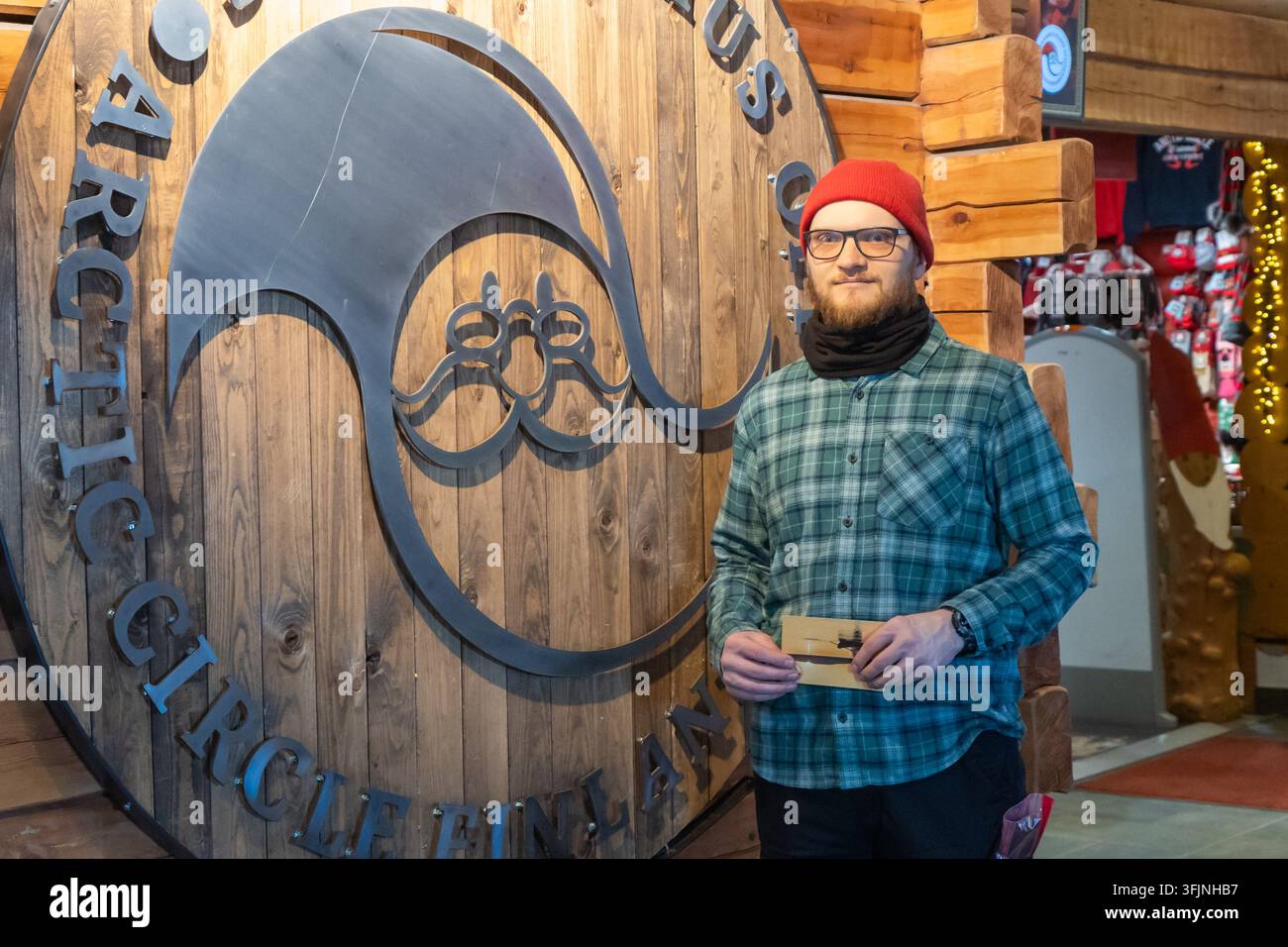 Rovaniemi, Finlandia - 15 maggio 2026: Un giovane con occhiali, un berretto rosso e una camicia a quadri sorride in piedi accanto a un grande sig decorativo in legno Foto Stock