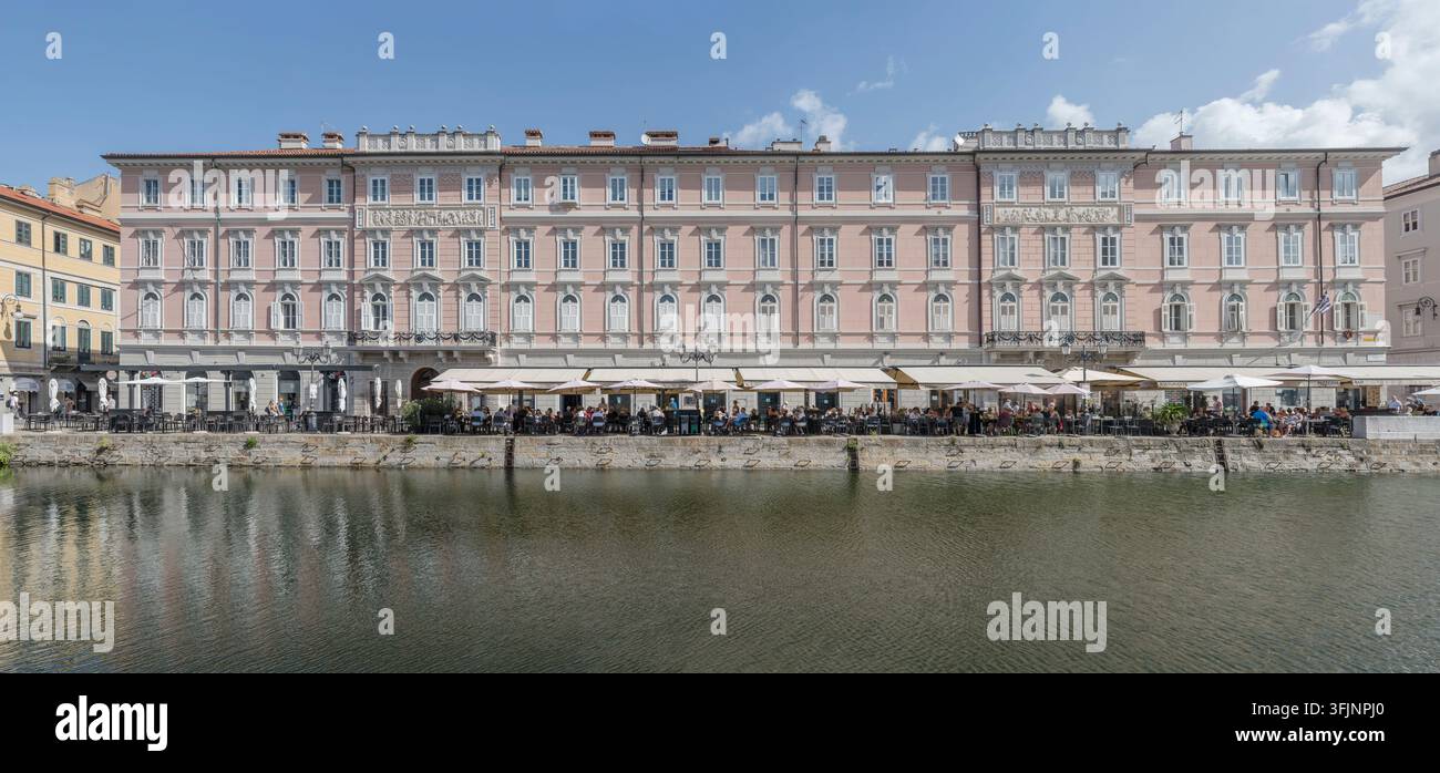 TRIESTE, 2026, 29 agosto; grande formato panorama editoriale della città con persone che si rilassano ai tavoli di luoghi pubblici sul lungotevere del Canal Grande sotto classe Foto Stock