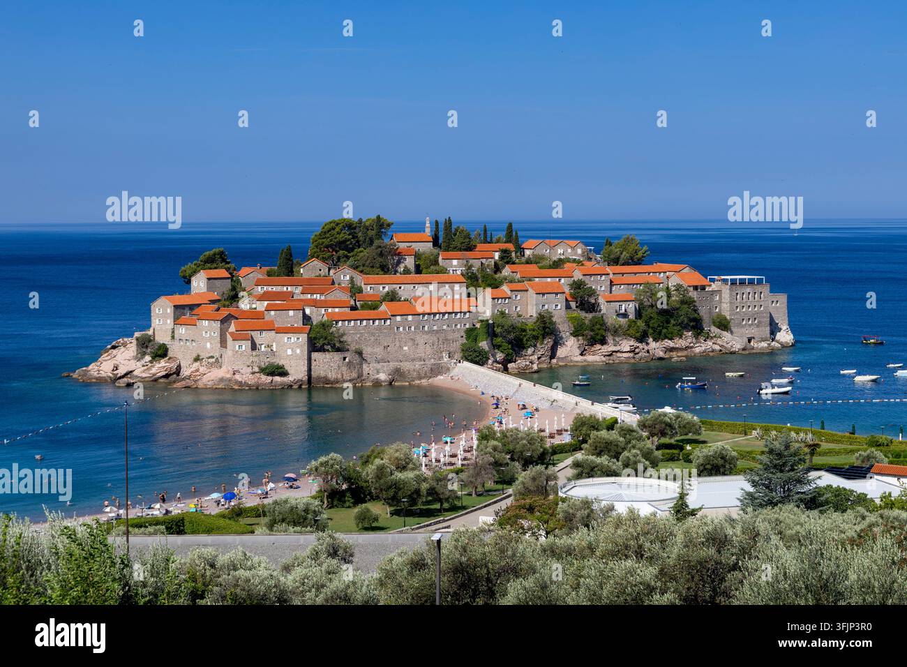 2 luglio 2026: Sveti Stefan, Montenegro, uno stretto pontile di pietra si estende sull'acqua costiera luminosa mentre i pedoni attraversano verso l'isola mediterranea fortificata Foto Stock