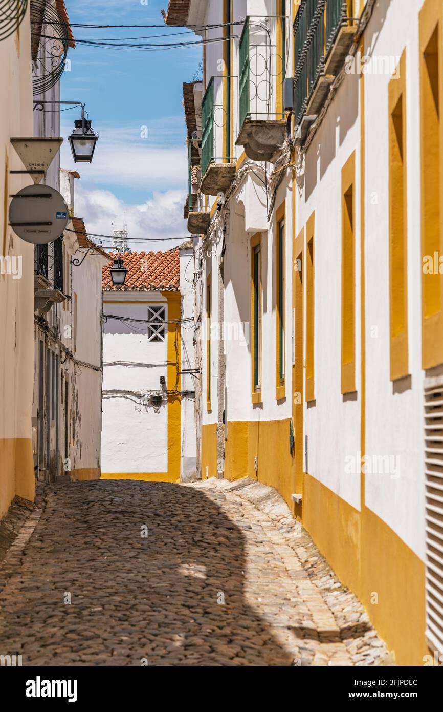 Vista di una stretta strada acciottolata nel centro storico di Évora, con edifici tradizionali imbiancati, rifiniture gialle e lanterne appese a Evo Foto Stock