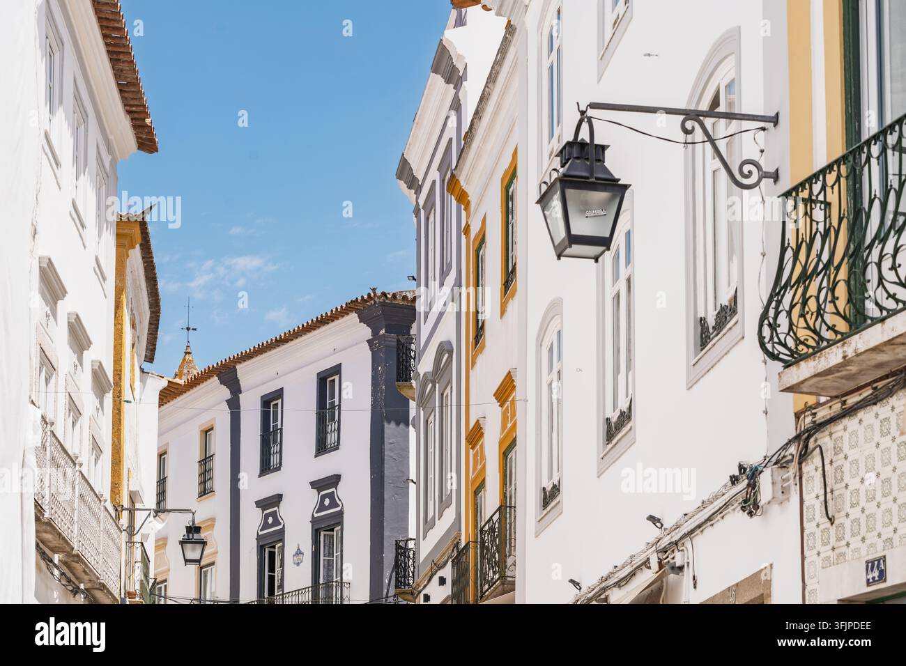 Vista di una strada storica a Évora con edifici bianchi, decorazioni gialle e grigie, balconi in ferro battuto e lanterne appese sotto un cielo blu terso a Évora Foto Stock