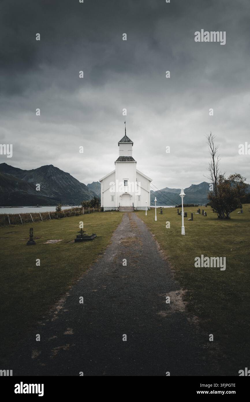 Vista di Gimsoy Kirke, una chiesa di legno bianca, situata alla fine di un sentiero di ghiaia in un cimitero erboso sotto un drammatico cielo nuvoloso a Gimsoysand, Nord Foto Stock