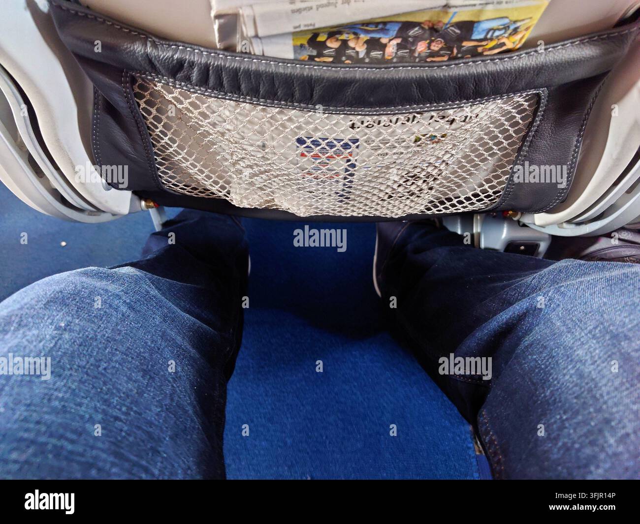 Vista ravvicinata dello spazio per le gambe di un passeggero in un aereo, mostra le gambe di una persona in jeans blu seduta comodamente. Lo schienale del sedile davanti mostra Foto Stock