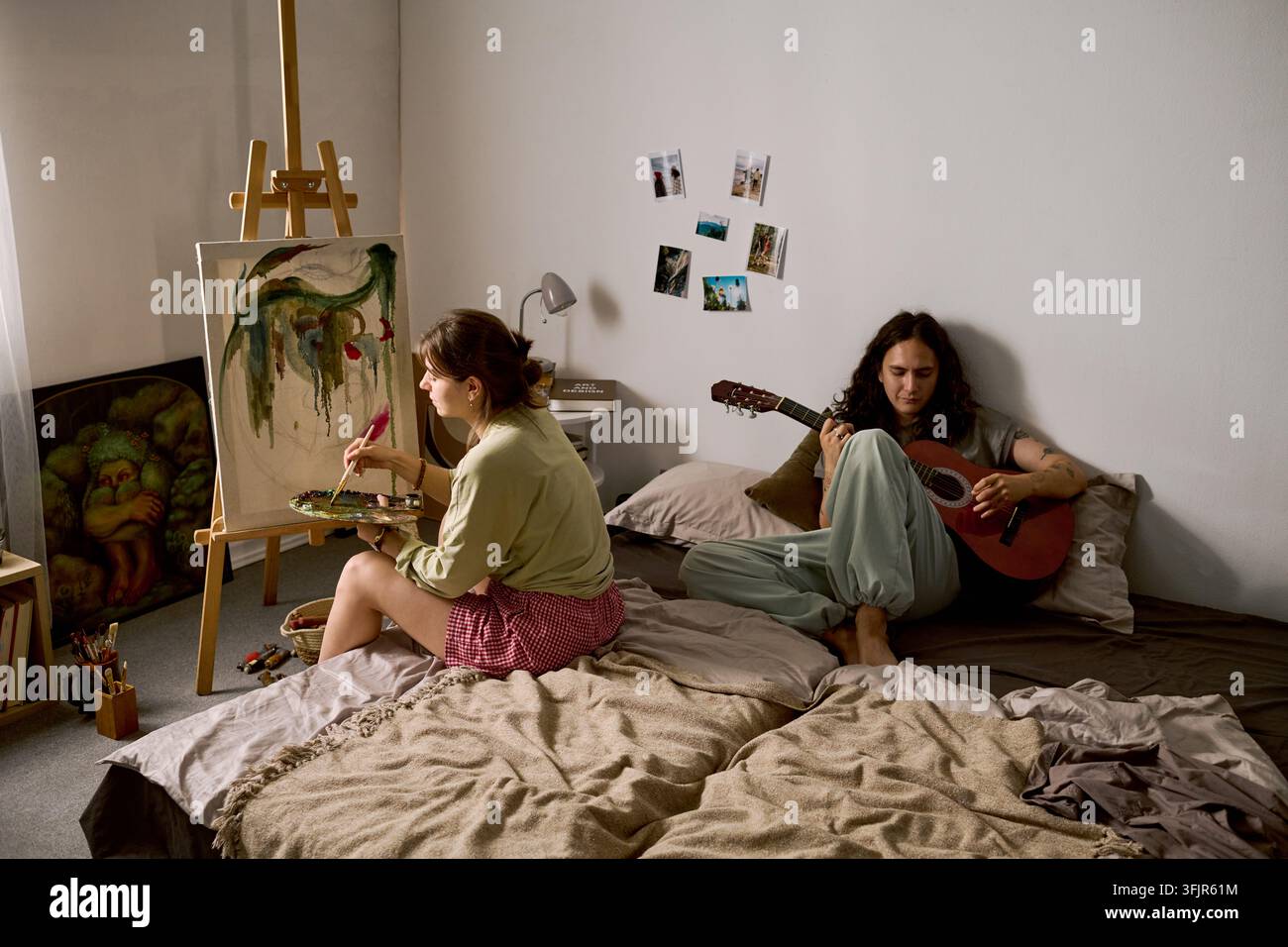 Donna che dipinge su tela mentre un uomo suona la chitarra acustica in camera da letto Foto Stock