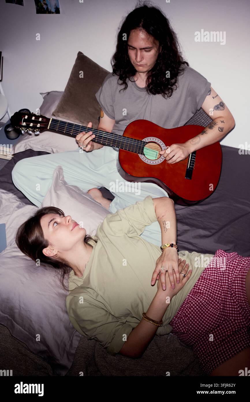Uomo che suona la chitarra acustica per una donna rilassata a letto a casa Foto Stock