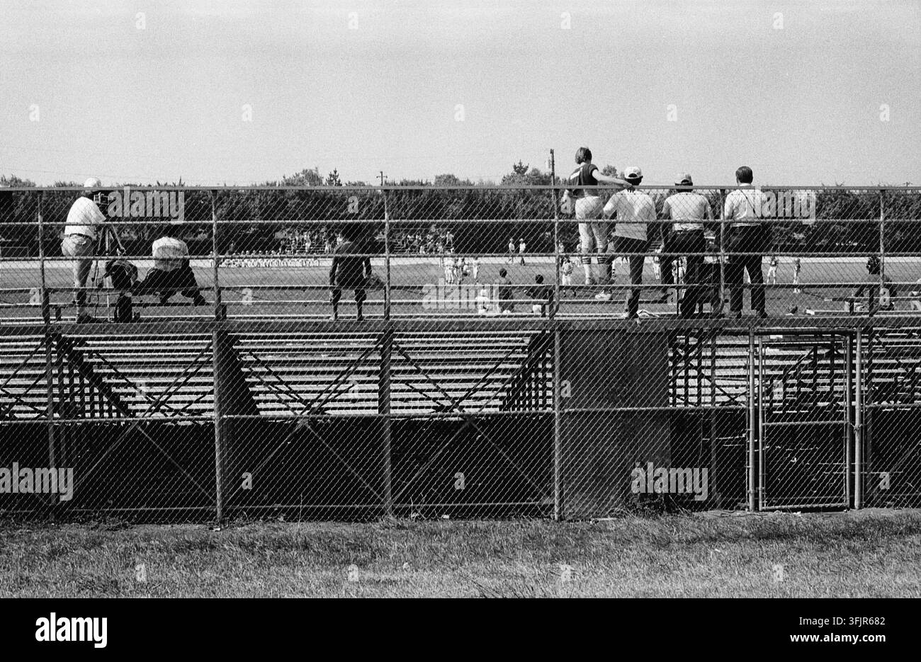 Catturata a Milpitas, California nel 1982, questa scena raffigura una partita di baseball locale in corso. Gli spettatori, principalmente uomini adulti, osservano i giocatori giovanili dalle gradinate dietro una recinzione in reti metalliche. Il paesaggio circostante presenta campi erbosi e alberi lontani, caratteristici delle aree ricreative all'aperto della Contea di Santa Clara. Milpitas, Contea di Santa Clara, California, USA Foto Stock