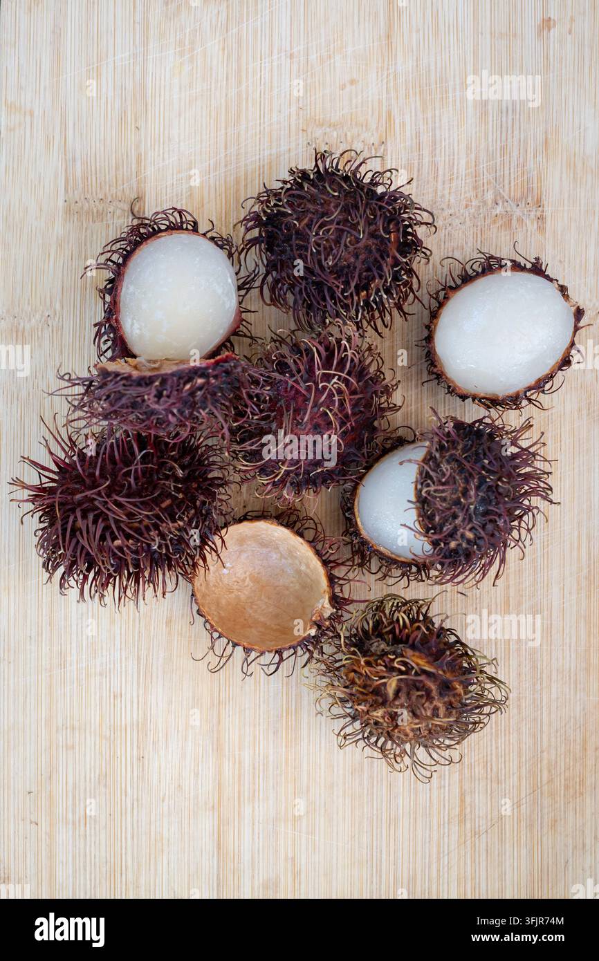 Flat lay di diversi frutti di rambutan aperti disposti su una superficie di legno rustico. La prospettiva dall'alto enfatizza la buccia spinosa rosso-marrone e il g Foto Stock