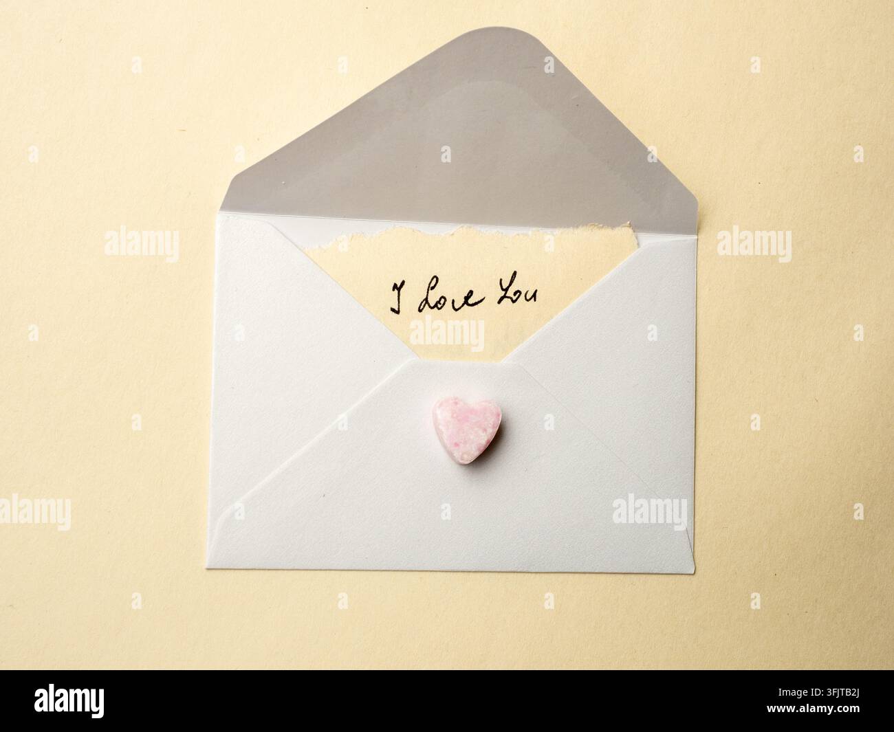 Una nota scritta a mano con 'Ti amo' spunta da una busta bianca sigillata con un piccolo cuore di caramella rosa, evocando romanticismo, affetto, nostalgia e intimità Foto Stock