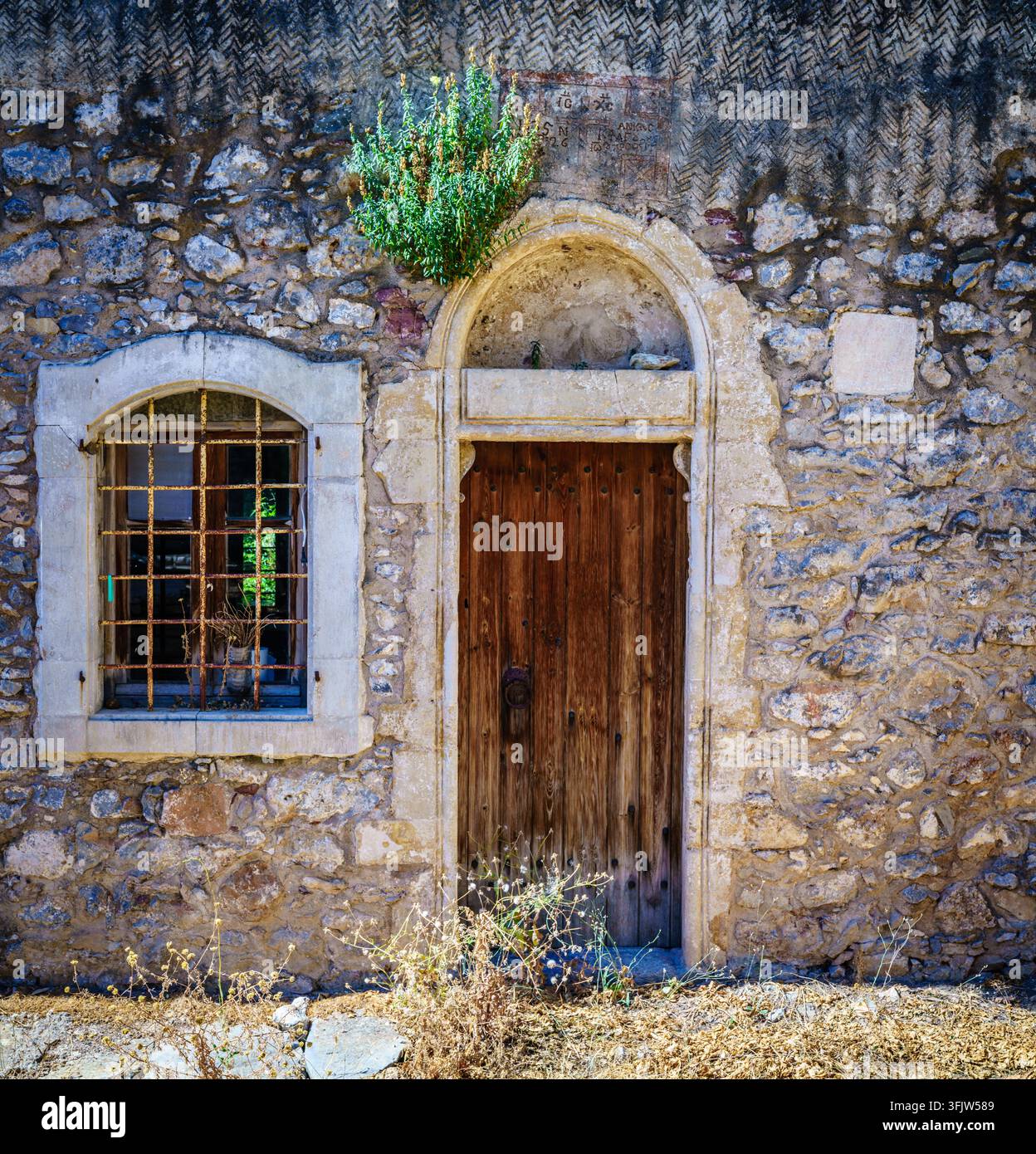 Primo piano della facciata di una casa tradizionale cretese con porta in legno invecchiata, finestra sbarrata e muro di pietra ruvido in un villaggio rurale Foto Stock