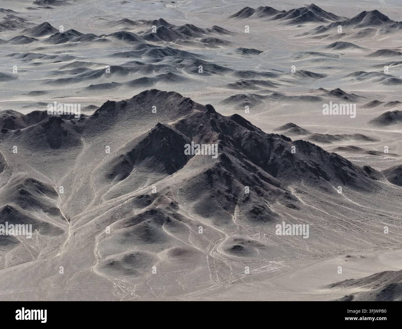 Un picco affilato e sterile si erge sopra pendici ricoperte di sabbia in questa veduta aerea di una catena desertica arida. Lievi tracce si snodano attraverso la natura selvaggia deserta sottostante. Foto Stock