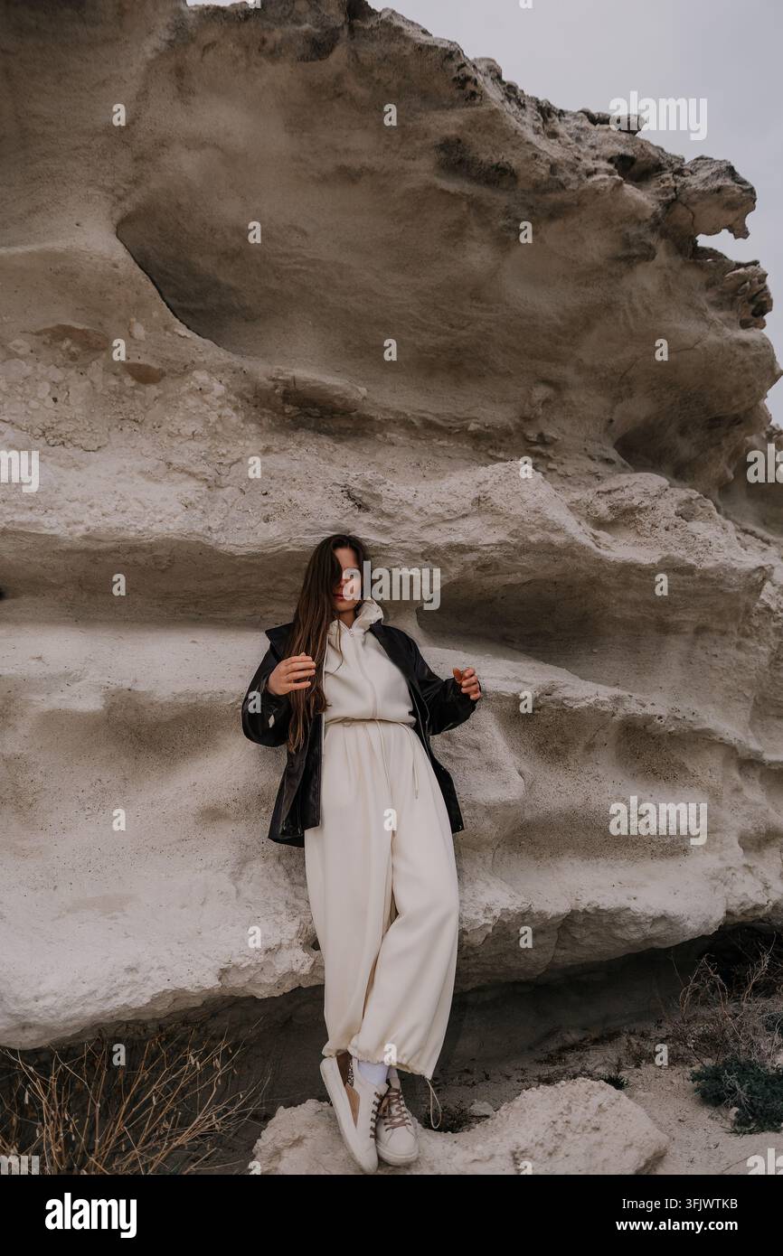 Donna in formazione rocciosa alla moda in piedi contro scogliere di pietra stratificate bianche che indossa una tuta bianca moderna e una giacca nera Foto Stock