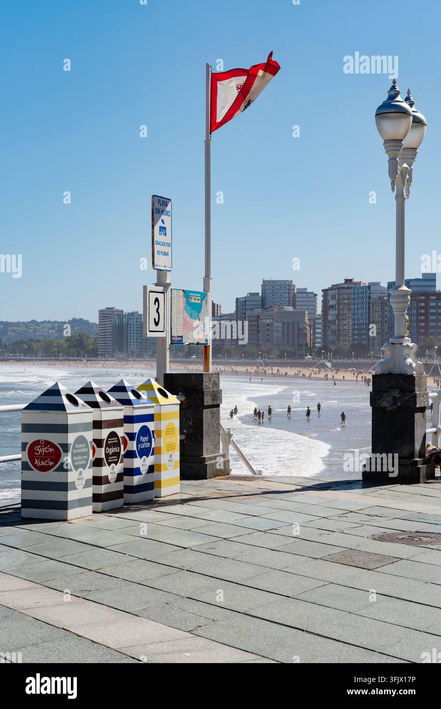 Gijon, Spagna – 19 settembre 2026 Contenitori di riciclo a strisce EMULSA allineano la passeggiata di San Lorenzo mentre i bagnanti si immergono nelle onde. Rifiuti municipali Foto Stock