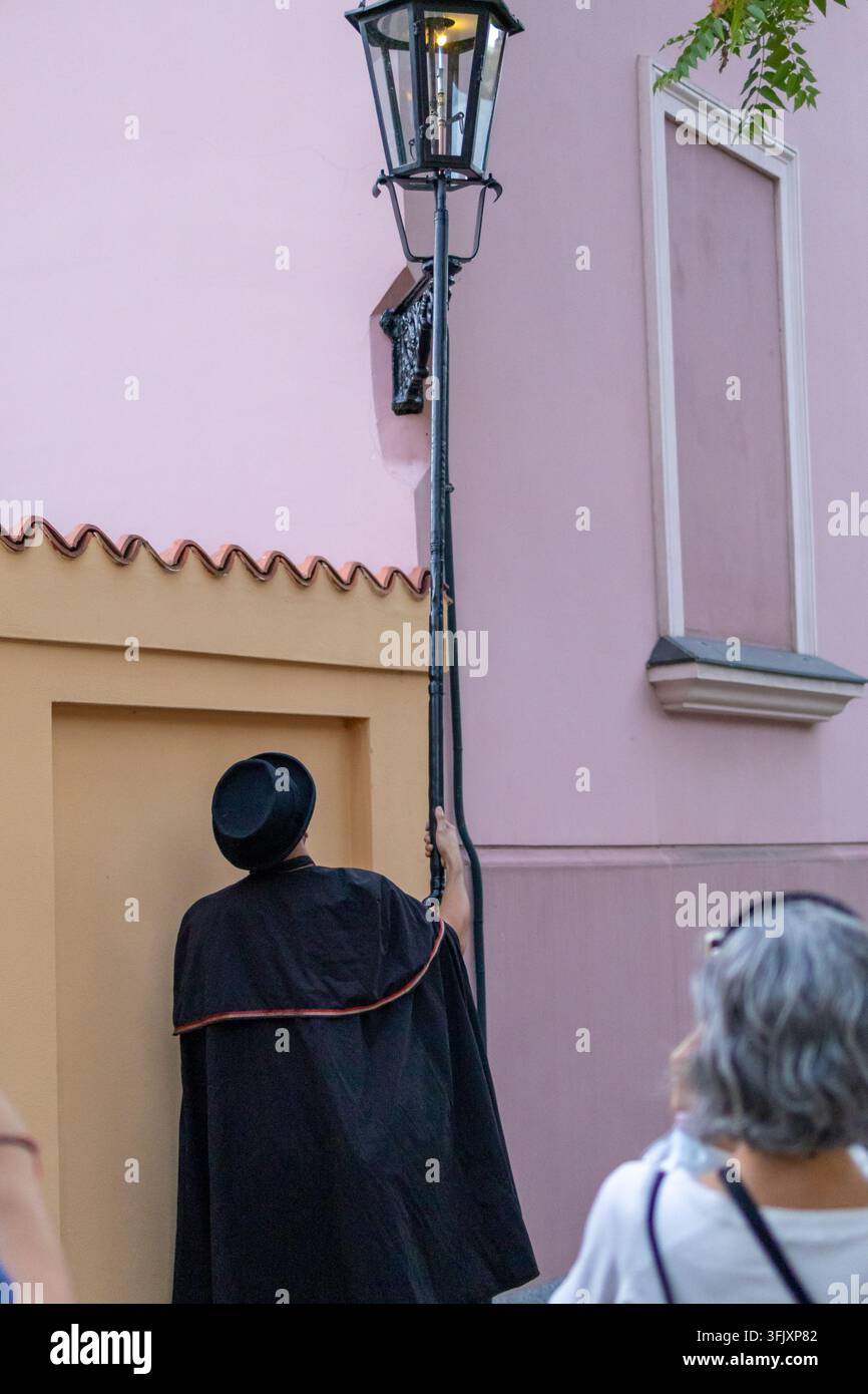 13 agosto 2026, Wroclaw, Polonia. Un tradizionale accenditore di lampioni usa una lunga asta per accendere una lanterna a gas vintage su un muro rosa mentre viene osservato dai turisti Foto Stock
