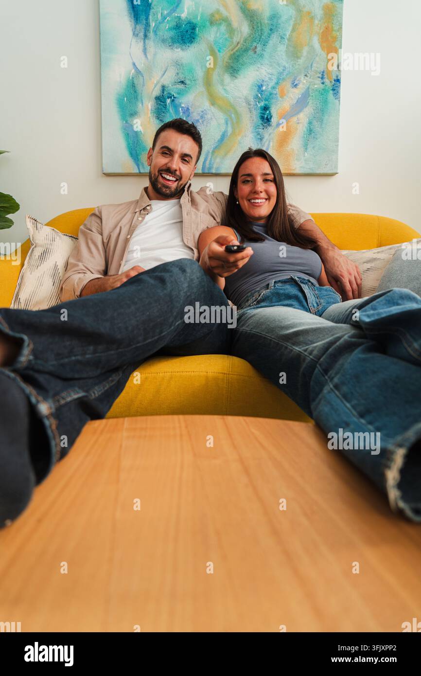 Una coppia allegra si rilassa su un divano giallo vivace, appoggiata indietro con sorrisi rilassati e compagnia calorosa, catturata per l'uso domestico dello stile di vita e Foto Stock