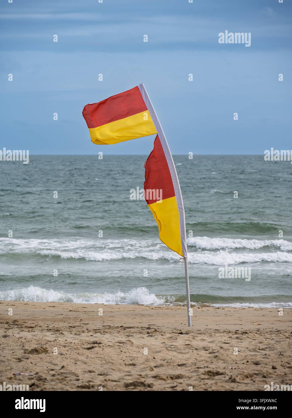 Una bandiera di salvataggio rossa e gialla che sventola nel vento su una riva di sabbia, segnando la zona di nuoto sicura contro le onde dell'oceano che si infrangono Foto Stock