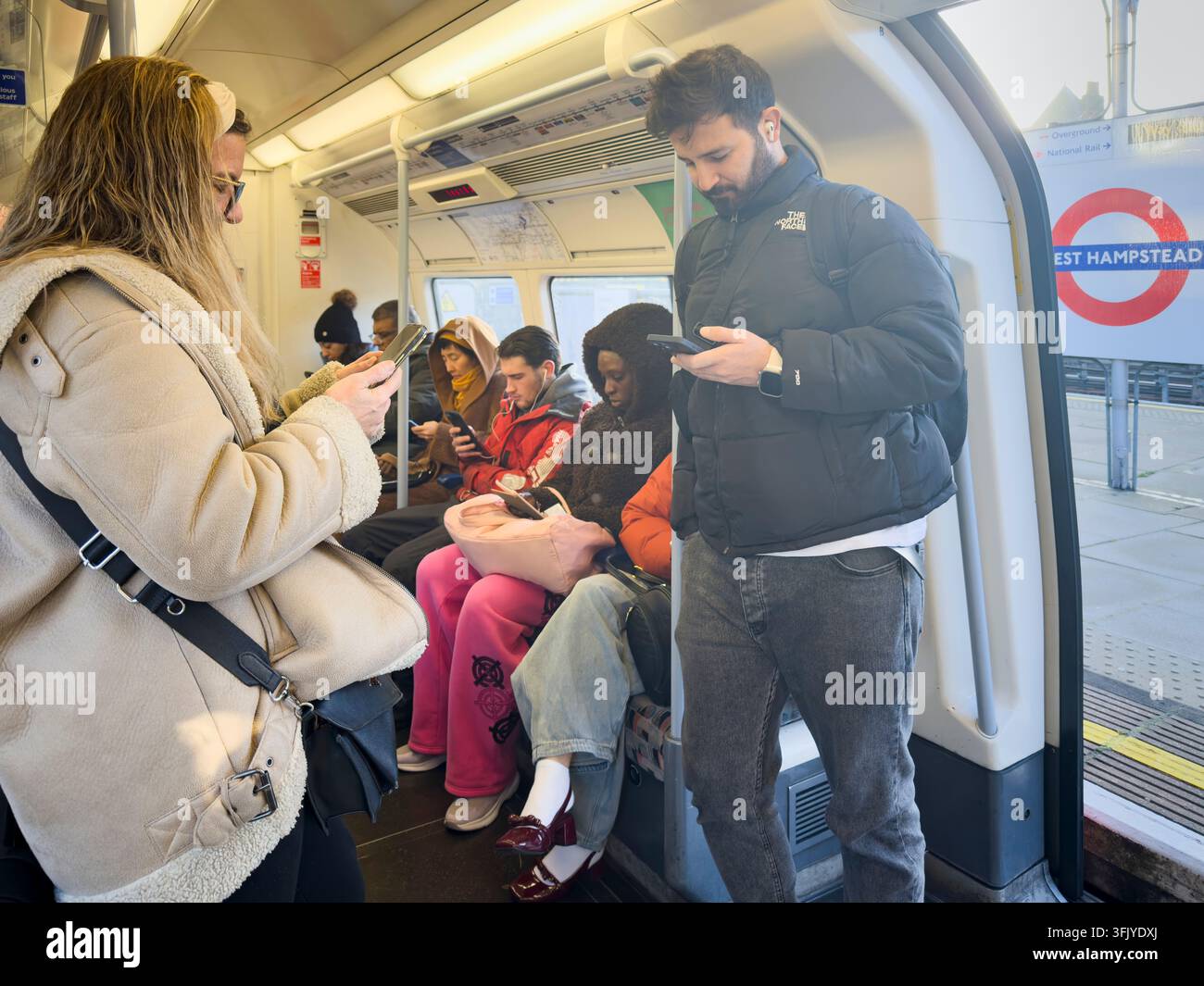I pendolari della metropolitana di Londra alla stazione di West Hampstead guardano i loro telefoni, passando il tempo durante il loro viaggio urbano quotidiano. Foto Stock