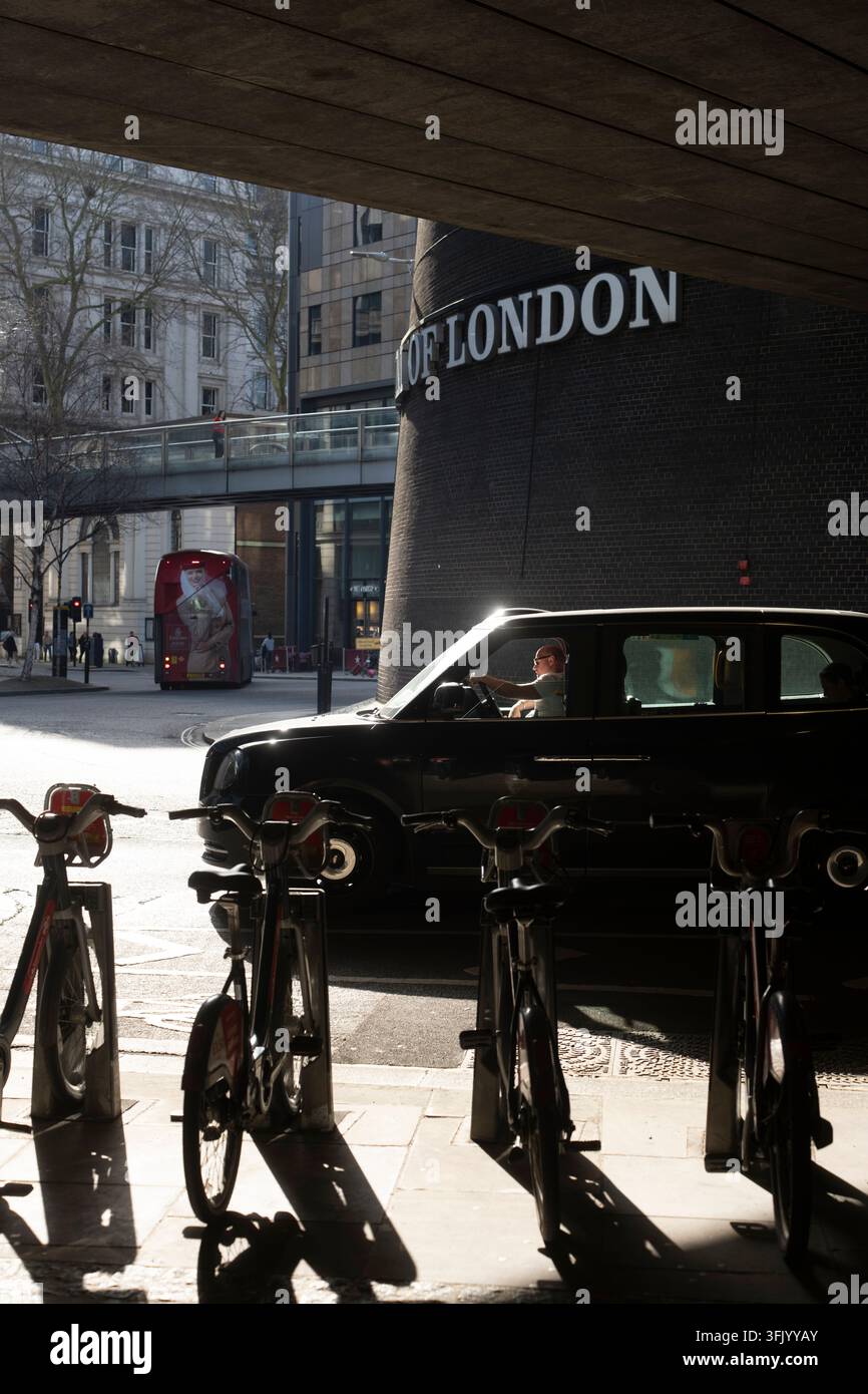 Un conducente guida un taxi nero oltre il sottopassaggio del Museo di Londra su London Wall, attraversando la città oltre biciclette parcheggiate. Foto Stock