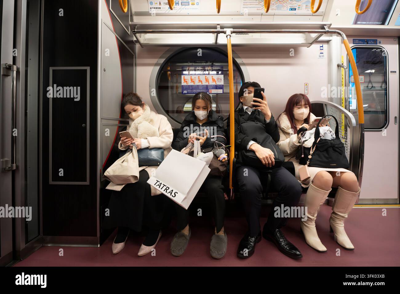 Quattro passeggeri mascherati controllano i loro telefoni mentre viaggiano su un treno della linea Marunouchi della Tokyo Metro con iconiche finestre circolari. Foto Stock