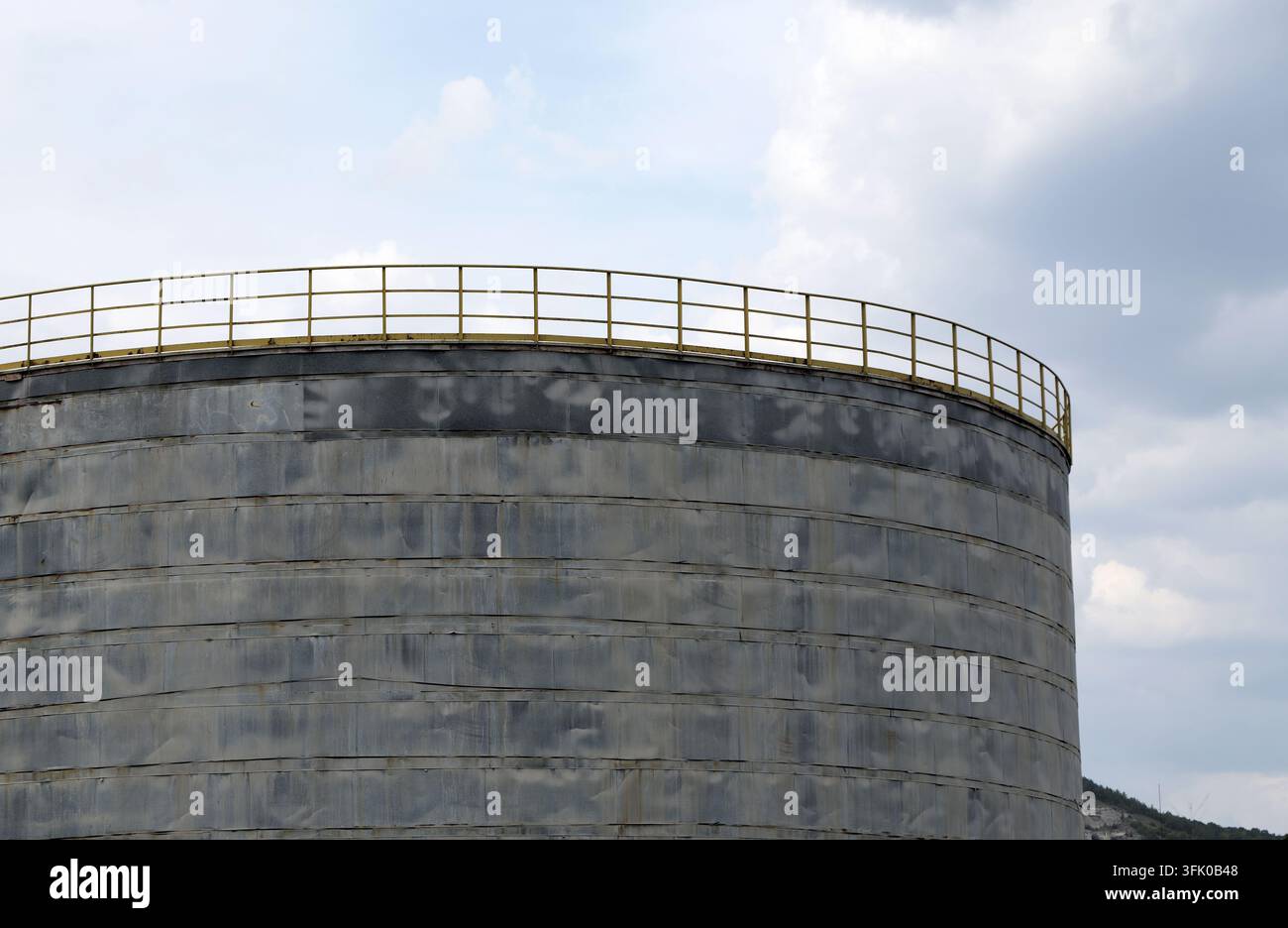 Grande serbatoio industriale di stoccaggio con una prominente ringhiera di sicurezza gialla sul perimetro superiore, in piedi sotto un cielo sereno con nuvole sparse, symb Foto Stock