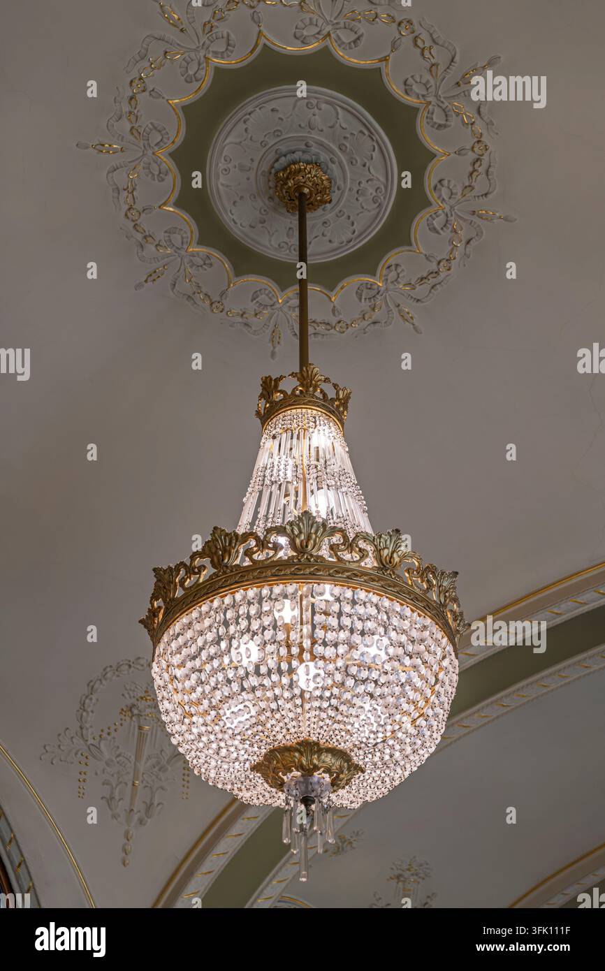 Un grande lampadario a cesto di cristallo pende sotto una rosetta di soffitto in gesso dorata circondata da modanature a ghirlanda, illuminando il soffitto a volta pallido di Foto Stock