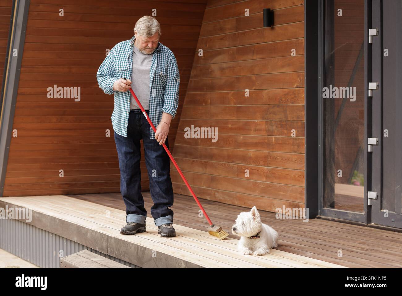 Un proprietario anziano spazza un portico di legno accanto al suo cane, illustrando la manutenzione esterna della casa, la compagnia dell'animale domestico e la vita quotidiana indipendente. Foto Stock