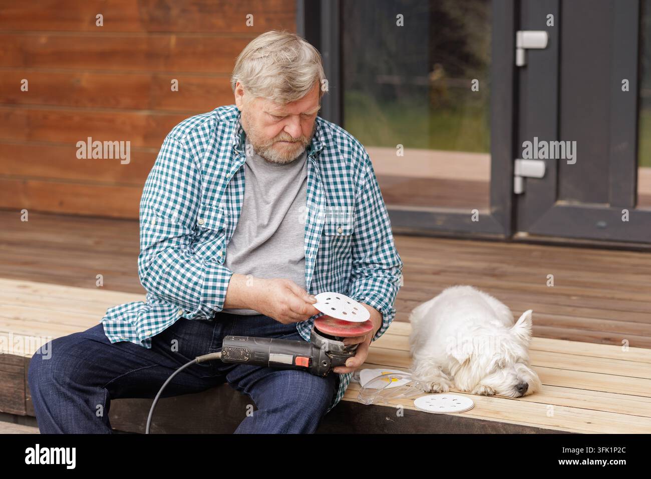 Un proprietario anziano utilizza una levigatrice orbitale per rifinire un terrazzo in legno all'aperto mentre il suo cane riposa nelle vicinanze, illustrando la manutenzione pratica della casa e Foto Stock