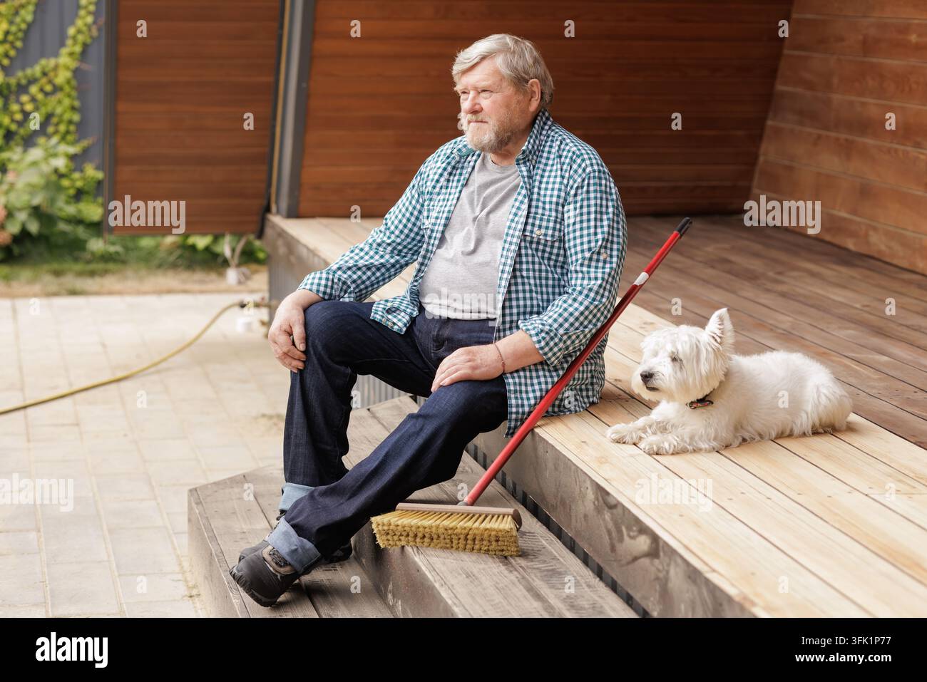Un uomo anziano è seduto accanto a una scopa su un portico di legno mentre il suo cane riposa nelle vicinanze, illustrando la cura della casa, la compagnia e una vita attiva in pensione. Foto Stock