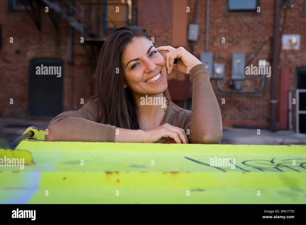Ritratto ravvicinato di una donna bruna che guarda l'obiettivo con una mano vicino alla fronte. Indossa un top di maglia marrone e jeans blu su uno sfondo vivace y Foto Stock