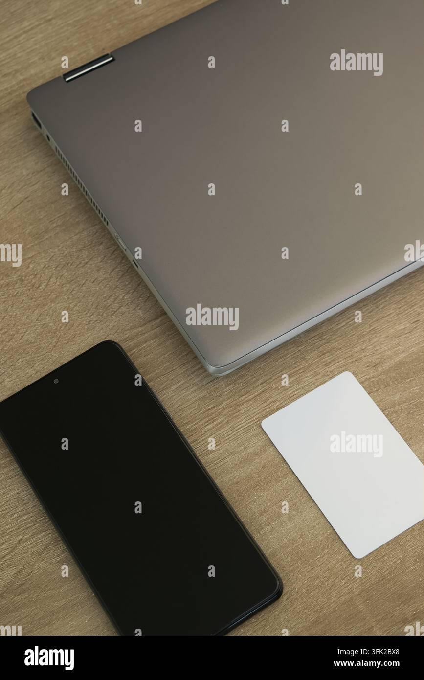Vista dall'alto di un laptop argentato chiuso, uno smartphone nero con schermo vuoto e una semplice carta d'accesso digitale bianca disposti su una superficie di scrivania in legno, rappresentante Foto Stock