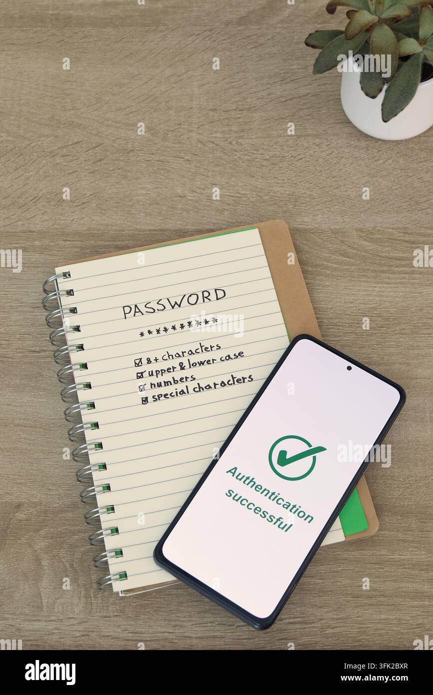 Smartphone che mostra il messaggio "Autenticazione riuscita" appoggiato diagonalmente su un notebook con una lista di controllo della forza della password, su una scrivania di legno accanto a un sm Foto Stock