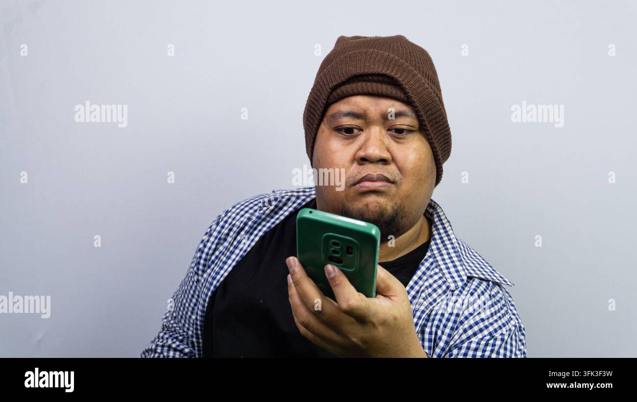 Un uomo asiatico serio con un berretto di lana marrone e una camicia a quadri fissa intensamente uno smartphone verde. L'individuo mostra una conce Foto Stock