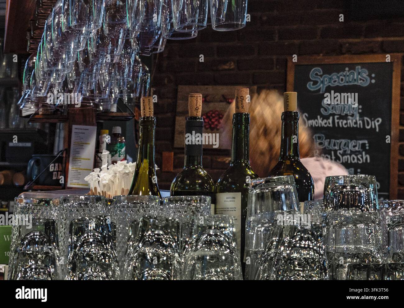 Un bancone da bar con vetreria trasparente impilata in primo piano, bottiglie di vino in piedi dietro, bicchieri a stelo appesi sopra e un cartello in lavagna sul Foto Stock