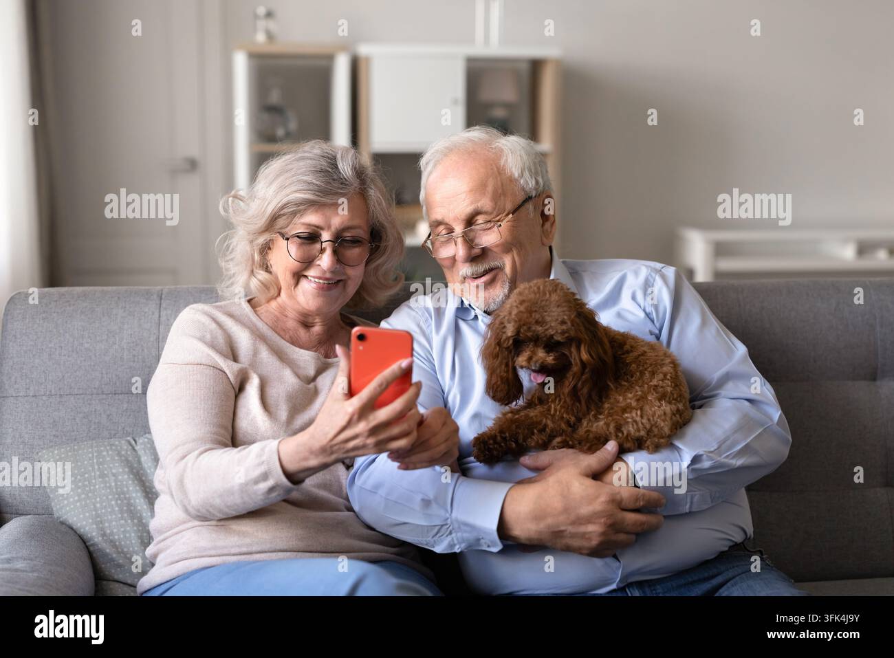 Coppia anziana che controlla lo smartphone, rilassata sul divano con un cane poodle Foto Stock