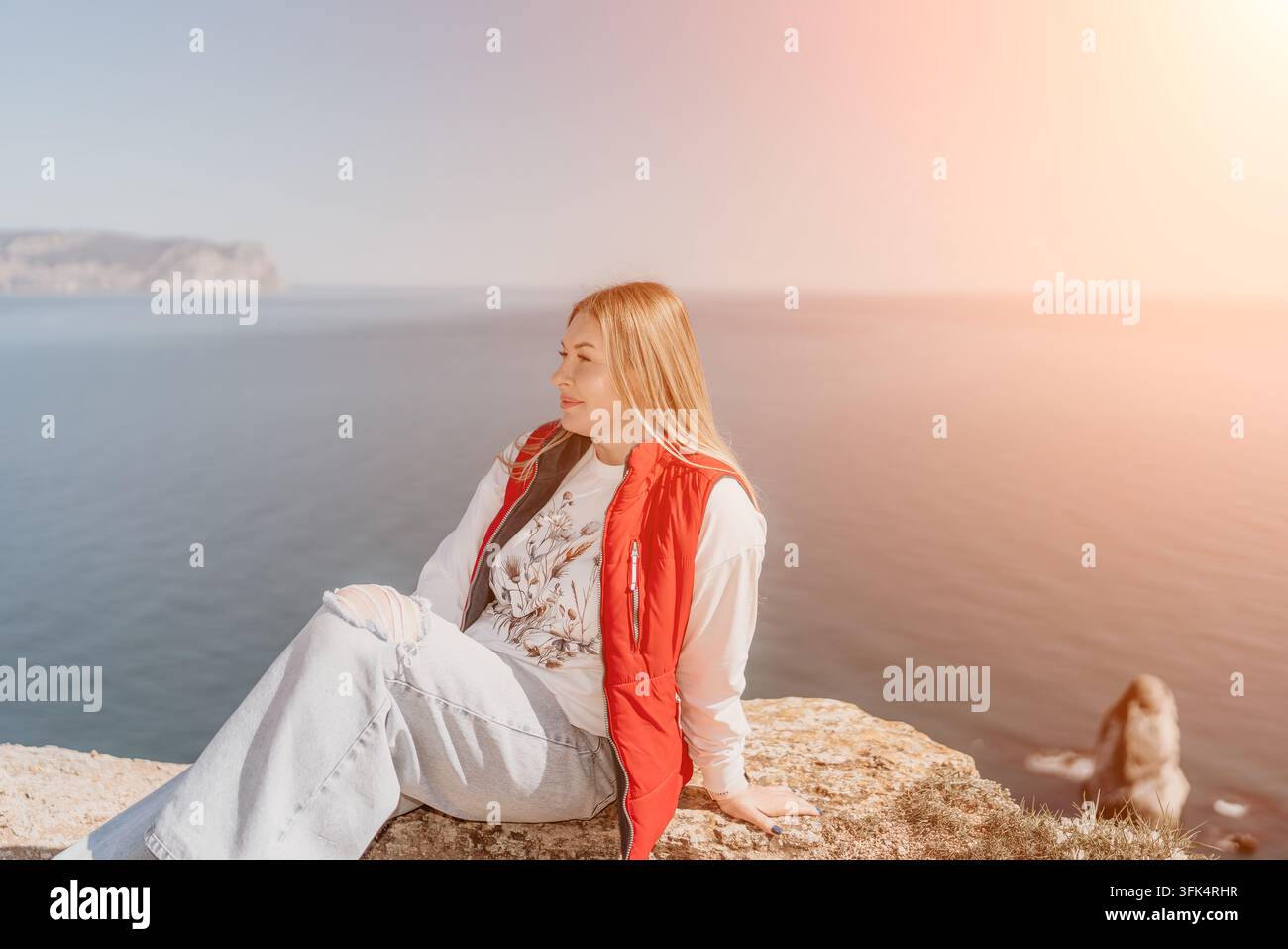 Donna sul litorale seduta sul bordo di una scogliera rocciosa che indossa un giubbotto rosso e si gode la pacifica vista sull'oceano Foto Stock