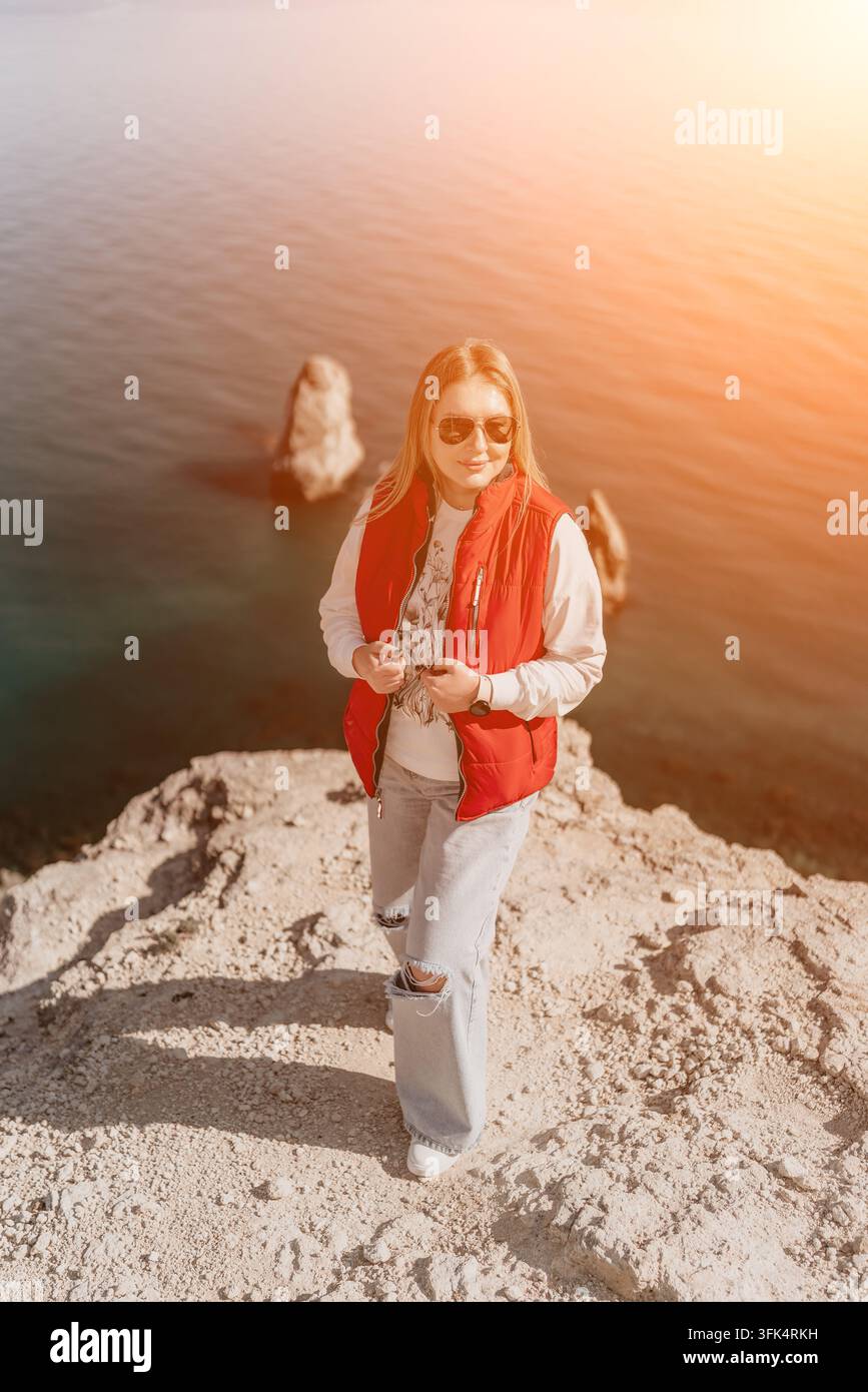 Donna sulla scogliera del mare si trova su una montagna rocciosa indossando un giubbotto rosso durante un tramonto luminoso che si affaccia sull'acqua Foto Stock