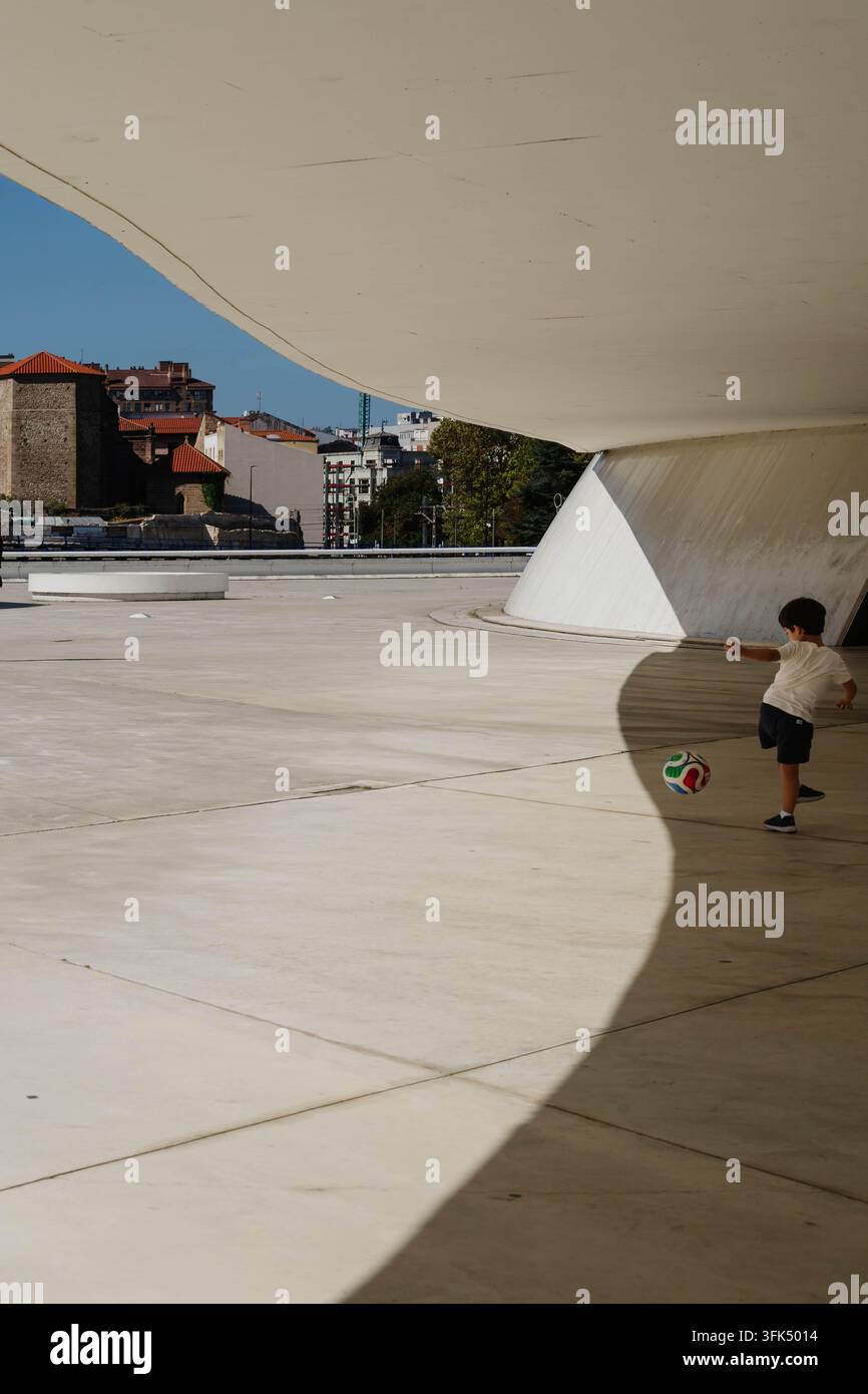 Aviles, Spagna - 20 settembre 2026 Un ragazzo calcia un pallone da calcio attraverso la piazza in cemento sotto l'auditorium bianco curvo del Centro Niemeyer. Urbano Foto Stock