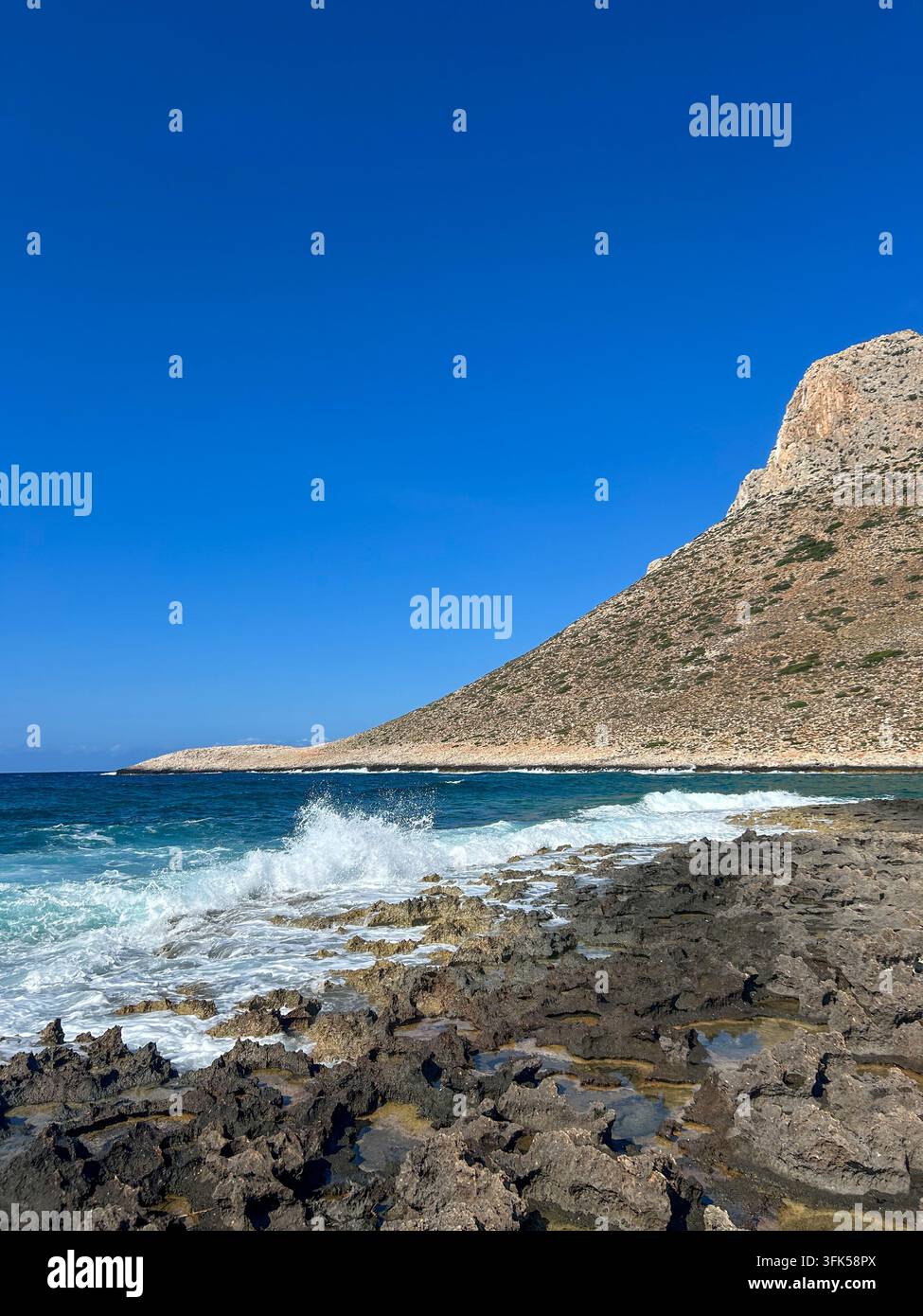 Onde potenti si infrangono su rocce vulcaniche frastagliate sotto un imponente picco montuoso sulla costa di Stavros, Creta, Grecia. - Immagine stock catturata con smartphone