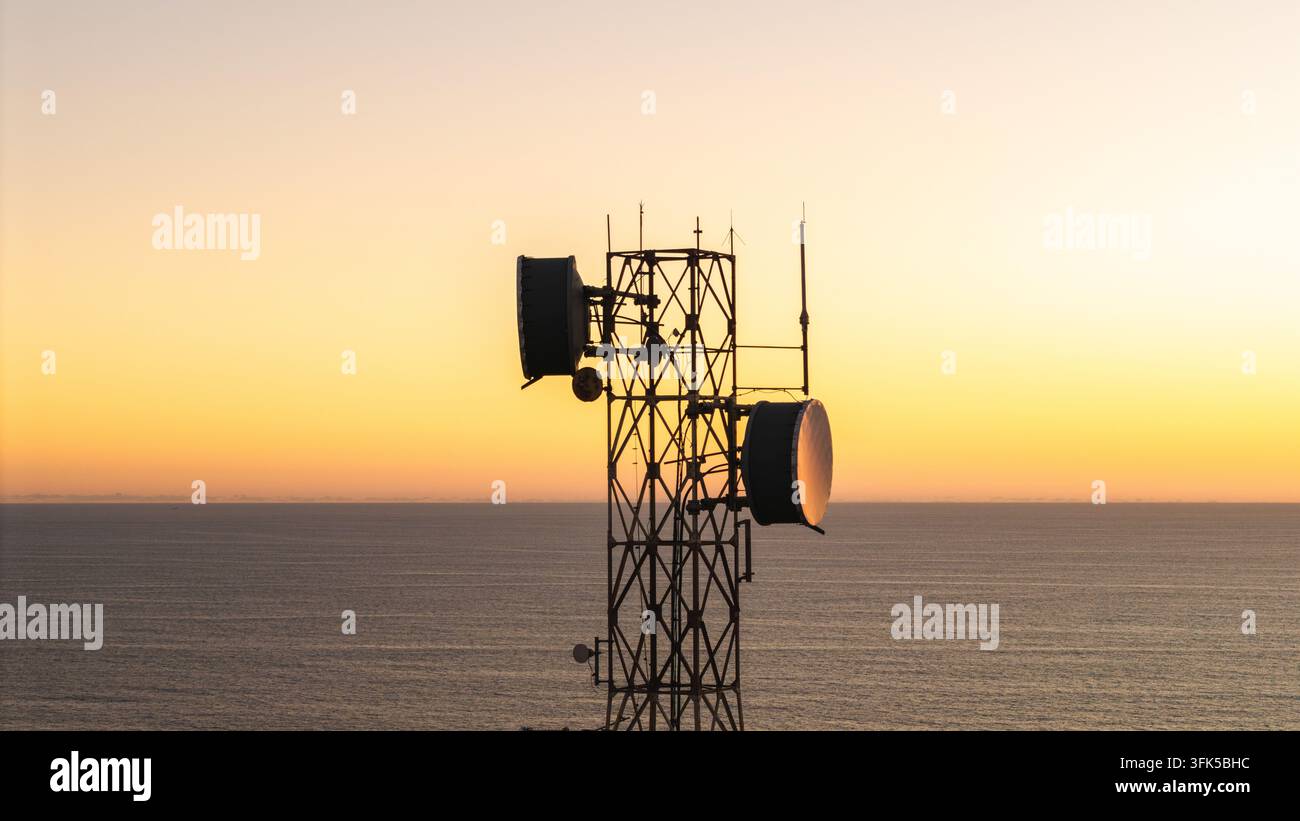 Torre di telecomunicazioni con parabole silouettate contro un tramonto luminoso sull'oceano. Foto Stock