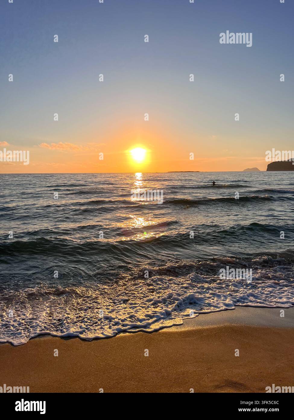 Un nuotatore solitario si gode il bagliore dorato del tramonto sulle onde gentili della spiaggia di Phalasarna, con la schiuma del mare che si riversa sulla sabbia, Creta. Foto Stock