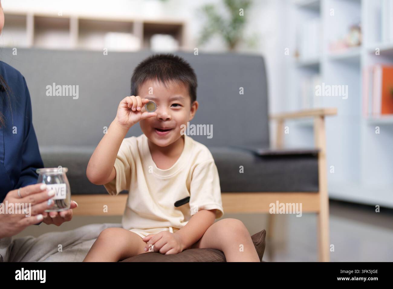 Ragazzo asiatico giocoso che guarda attraverso una moneta per divertimento, risparmio e educazione finanziaria Foto Stock