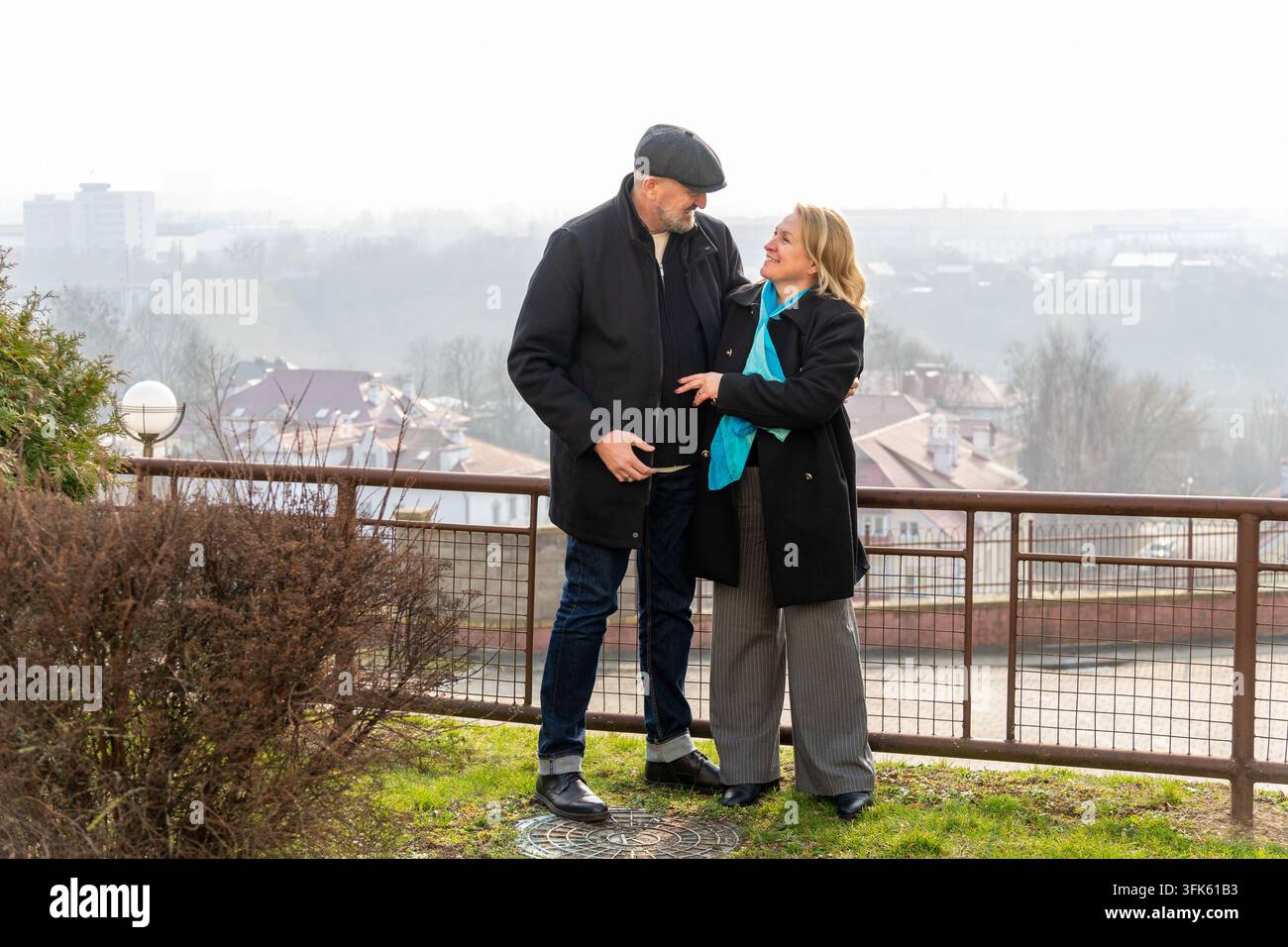 Coppia matura che si sorride su un punto di vista nebbioso della città, uomo con berretto piatto e donna con sciarpa blu in piedi vicini Foto Stock