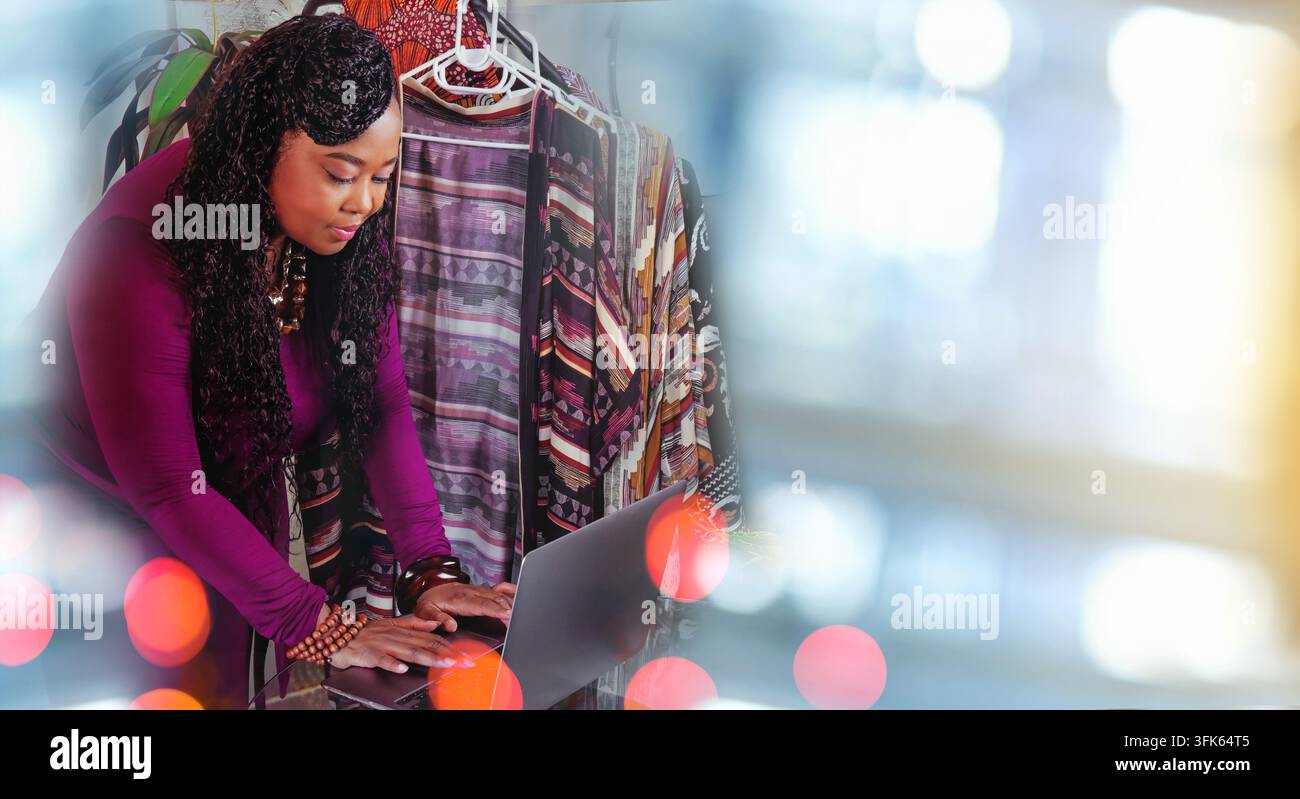 una stilista che rivede abiti a fantasia mentre lavora su un laptop nel suo studio creativo, con luci decorative a bokeh Foto Stock