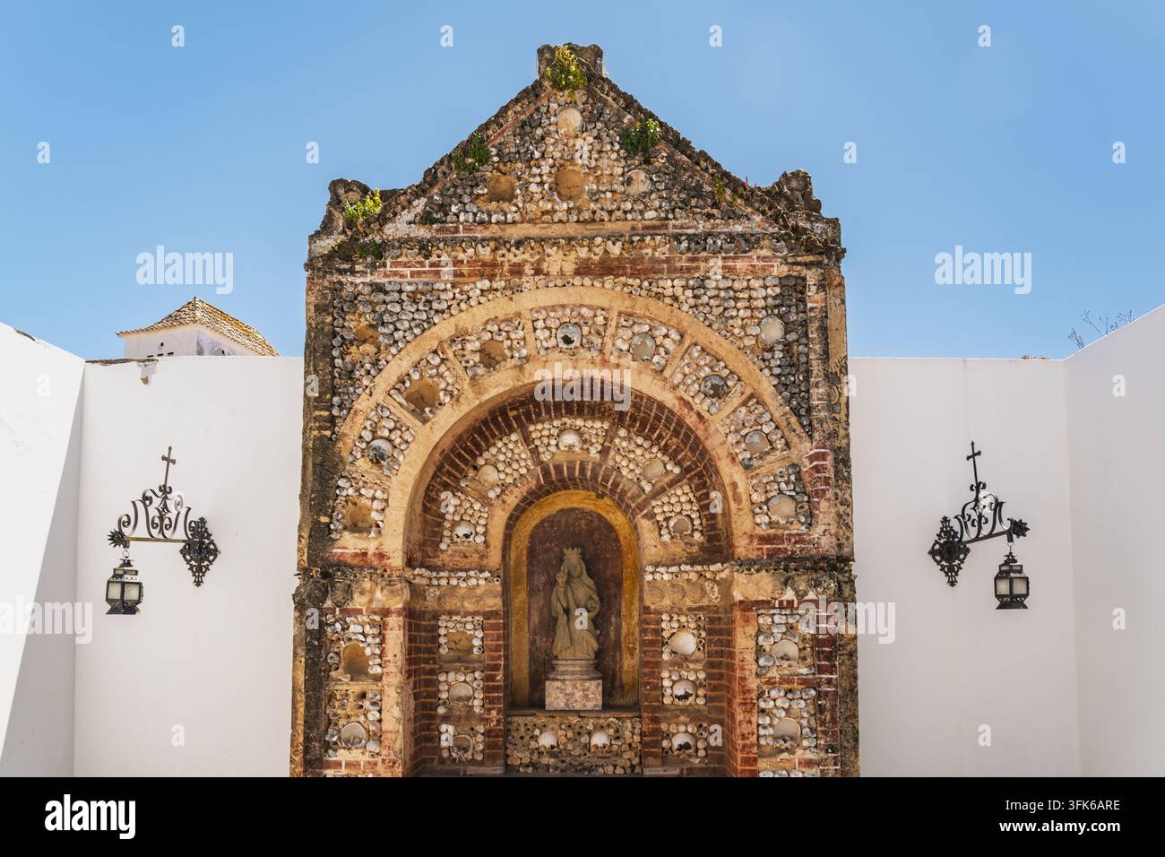 Vista dell'ingresso ornato della Cappella delle Ossa decorata con conchiglie e ossa, con una statua centrale e lanterne in ferro battuto su uno sfondo blu Foto Stock