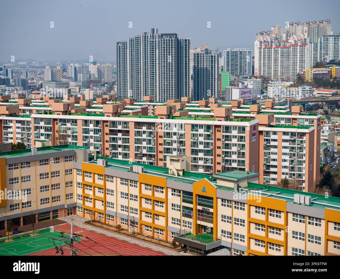Busan, Corea del Sud - 07 febbraio 2026: Vista di un moderno edificio scolastico con una facciata gialla e arancione e un tetto verde, situato di fronte a una zona residenziale densamente popolata Foto Stock
