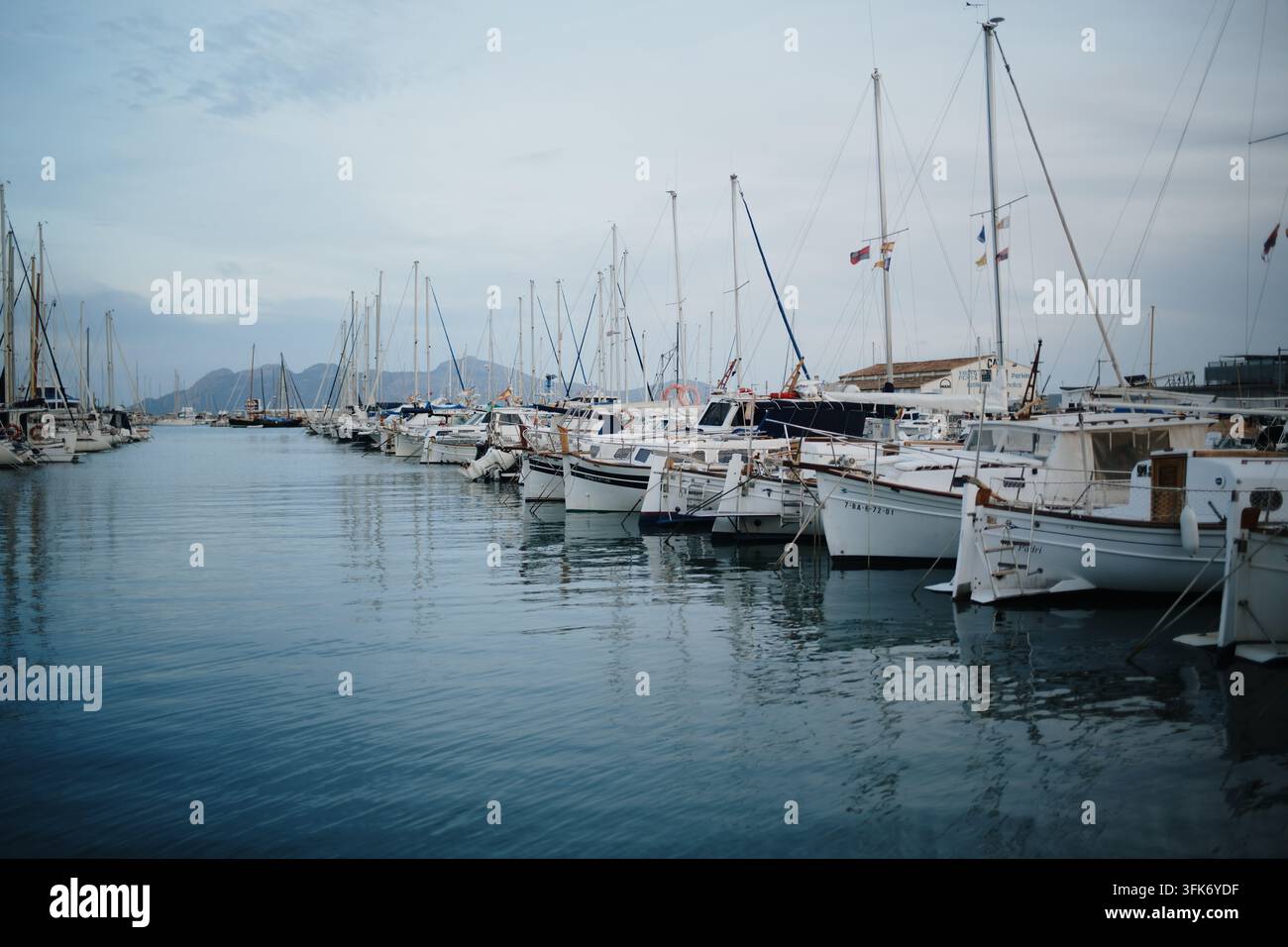 Port de Pollenca, Spagna – 05 gennaio 2025: Vista di barche a vela e yacht ormeggiati nel porto turistico con acqua calma e colline lontane sotto un cielo nuvoloso. Foto Stock