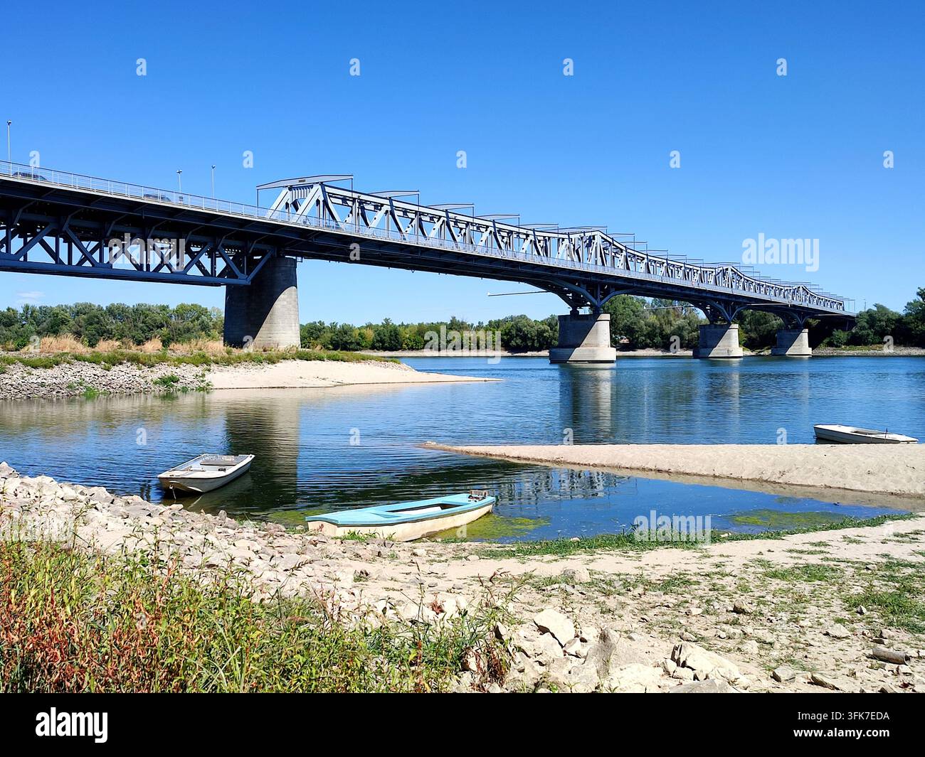 Vista ampia del ponte sul Danubio a Baja, Ungheria, con piccole barche che riposano vicino a una riva sabbiosa sotto un cielo estivo blu e sereno. - Immagine stock catturata con smartphone