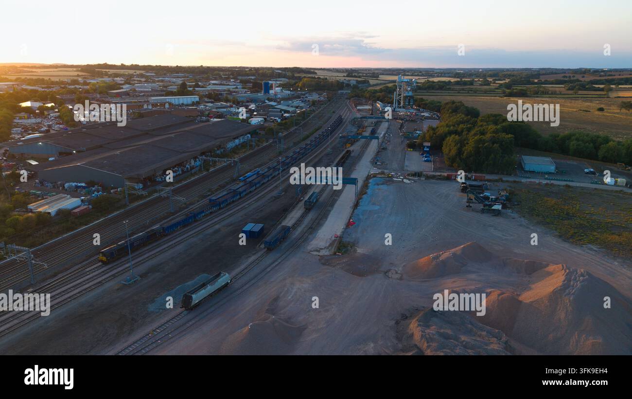 Vista aerea del terminal merci di Wellingborough al tramonto con treni merci sui binari, magazzini industriali e cumuli di inerti a Welli Foto Stock