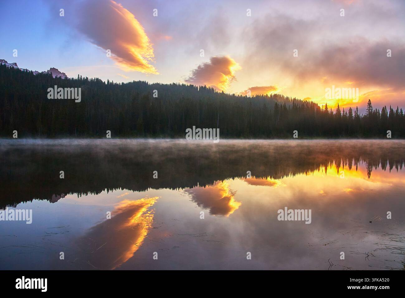 La luce soffusa del mattino e le nuvole colorate si riflettono sulle acque calme del Reflection Lake nello stato di Washington. La nebbia si alza sopra il lago mentre l'alba lo illumina Foto Stock