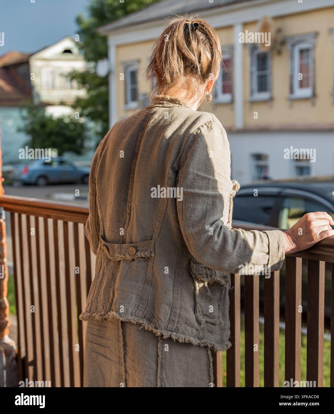 Donna si appoggia su una ringhiera di legno, di spalle alla macchina fotografica. I suoi capelli sono raccolti, la giacca ha i bordi sfilacciati. Una luce soffusa illumina la sua postura rilassata Foto Stock