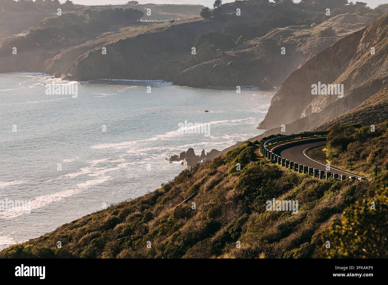Una strada curva con guardrail si snoda lungo ripide colline verdi e scogliere sopra l'oceano e insenature rocciose nella morbida luce crepuscolare nebbiosa. Foto Stock